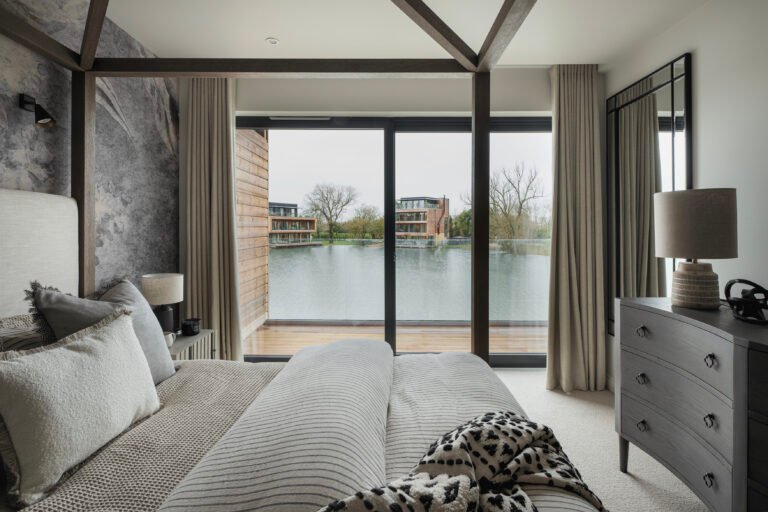 Serene bedroom with layered neutral textiles, a four-poster bed, and large windows overlooking a calm lakeside view