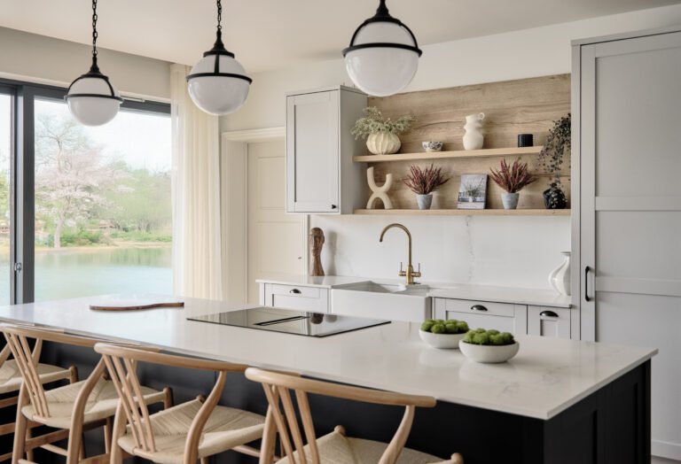 Luxury kitchen interior design in Bristol featuring a marble island, brass tap, open oak shelving and soft neutral cabinetry overlooking a lakeside view.